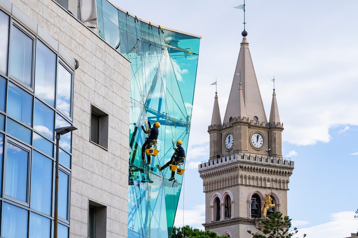 Dos operarios en suspensión limpian un muro cortina de vidrio en la fachada de un edificio moderno.